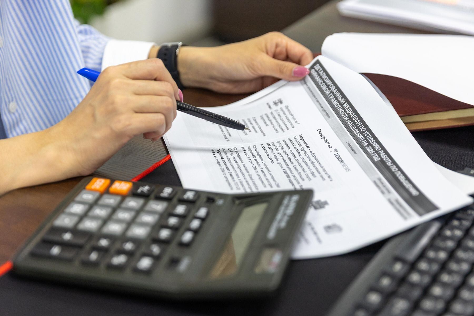 Mão segurando uma caneta sobre relatório contábil, ao lado de uma calculadora em uma mesa escura